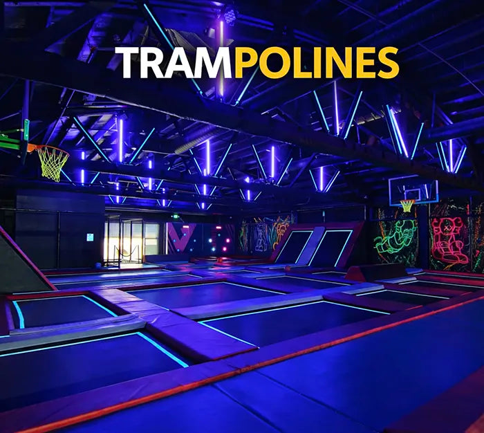 A neon-lit indoor trampoline park with basketball hoops, glowing trampolines, and vibrant wall art, under a dark ceiling with purple and blue lights. The word "TRAMPOLINES" is displayed at the top.