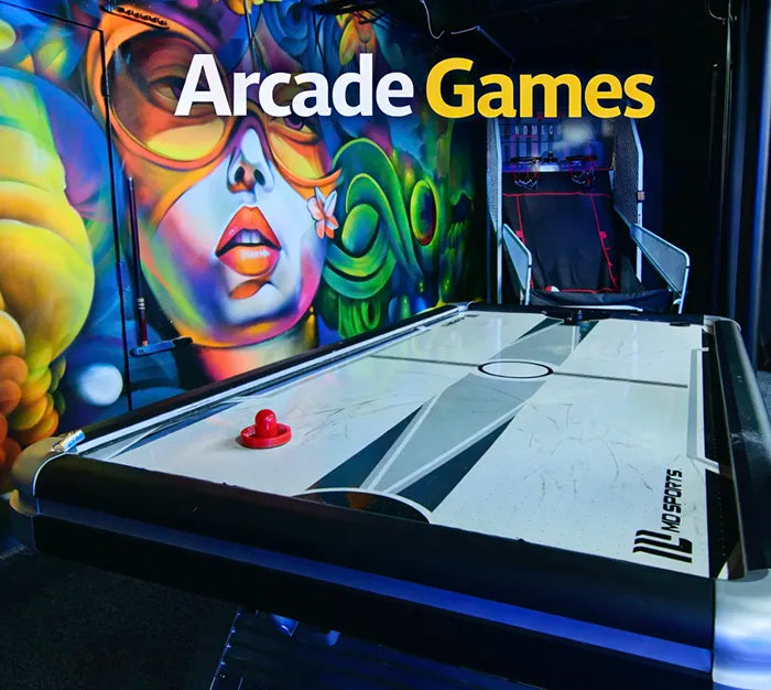 An air hockey table in front of a colorful graffiti mural featuring a woman's face and mask, with the words "Arcade Games" in large letters at the top.