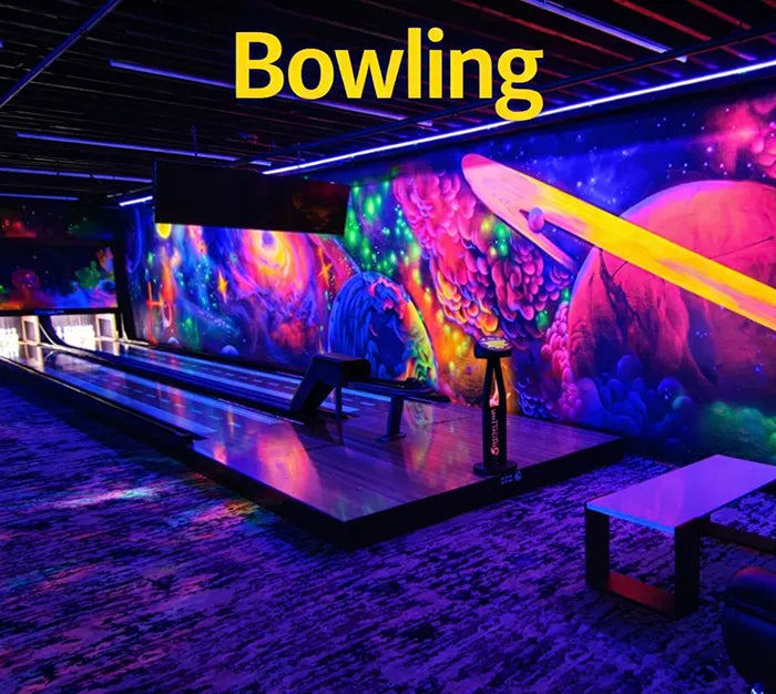 A colorful, neon-lit bowling alley with cosmic-themed wall art showing planets and galaxies. “Bowling” is written in large yellow text at the top of the image. The lanes and seating area glow under black lights.