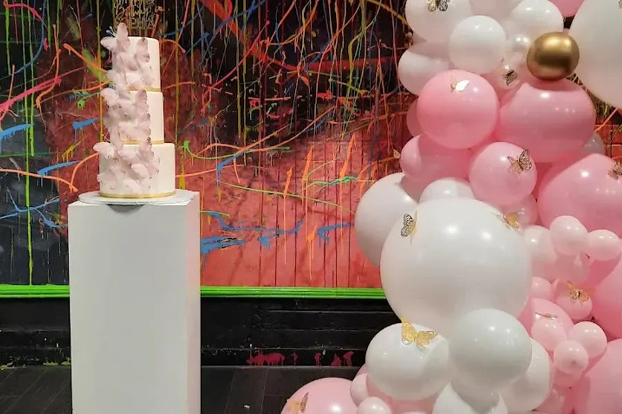 A tall, pink and white tiered cake on a pedestal stands beside a cluster of pink, white, and gold balloons, with a colorful, splatter-painted wall in the background.