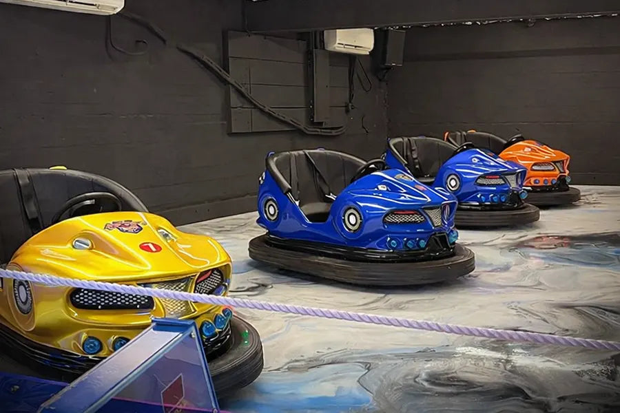 Four colorful bumper cars—yellow, blue, and orange—are parked indoors on a smooth, marbled floor behind a rope barrier. The room has dark walls and air conditioning units mounted above.