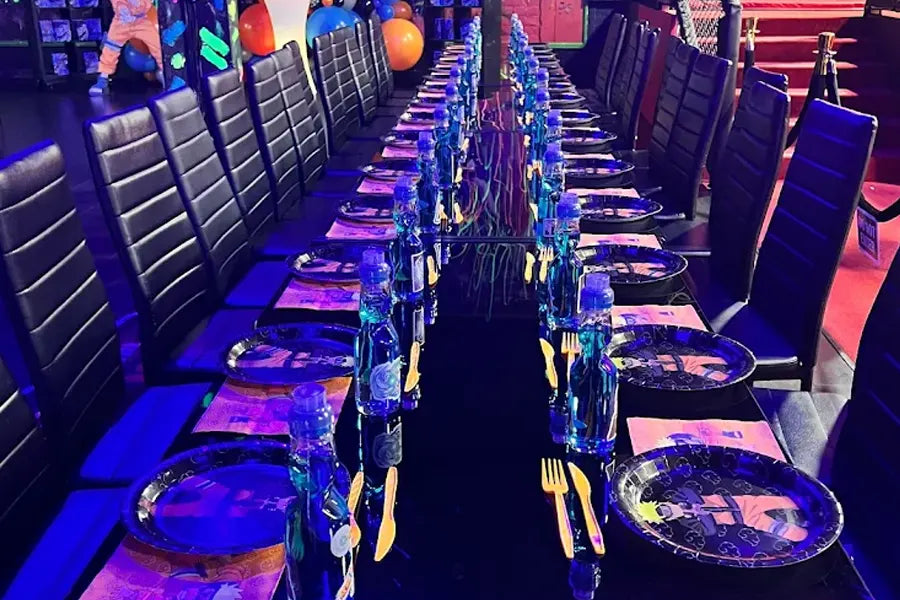 A long, black dining table is set for a party with black chairs, blue water bottles, black plates, and gold plastic cutlery. Colorful balloons and decorations are visible in the background under purple lighting.