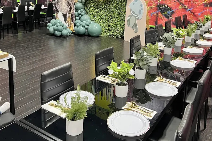 A modern dining area with black chairs and glossy tables set with white plates, gold cutlery, and small potted green plants. Decorative balloons and colorful wall art are in the background.