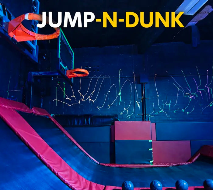 Indoor trampoline basketball court with padded ramps and walls, featuring two orange basketball hoops. The words “JUMP-N-DUNK” in bold white and yellow letters are at the top. Neon designs decorate the dark wall.