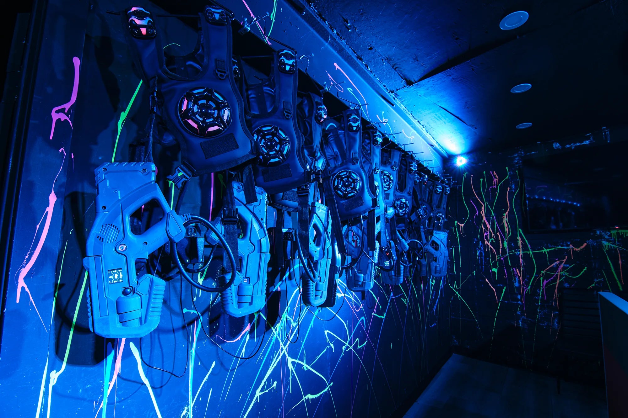 A row of laser tag vests and guns hangs on a dark wall illuminated by blue and neon lights, with colorful paint splatters decorating the background.