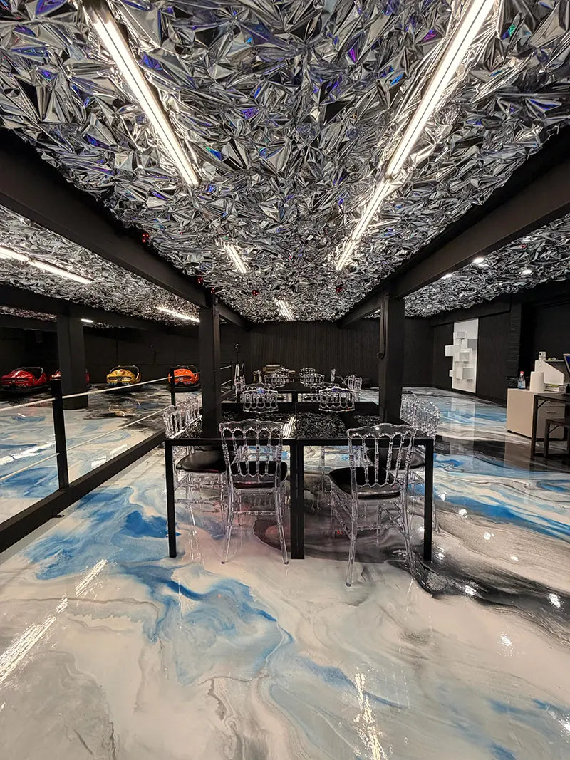 A modern room with shiny, reflective silver ceiling panels, clear chairs around black tables, and a glossy blue-and-white marbled floor; colorful sports cars are visible behind a glass wall.