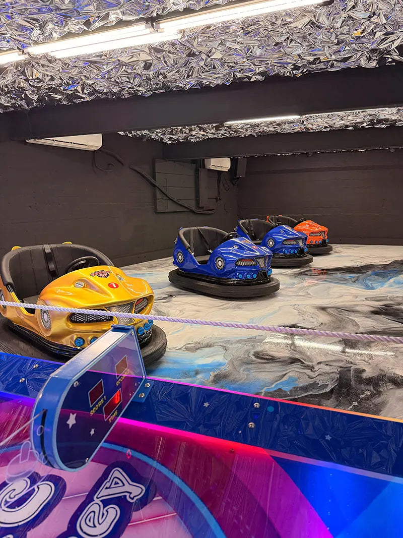Three bumper cars—yellow, blue, and orange—sit on a shiny, marbled indoor arena under a ceiling with reflective, metallic decorations. The setting appears empty and well-lit.