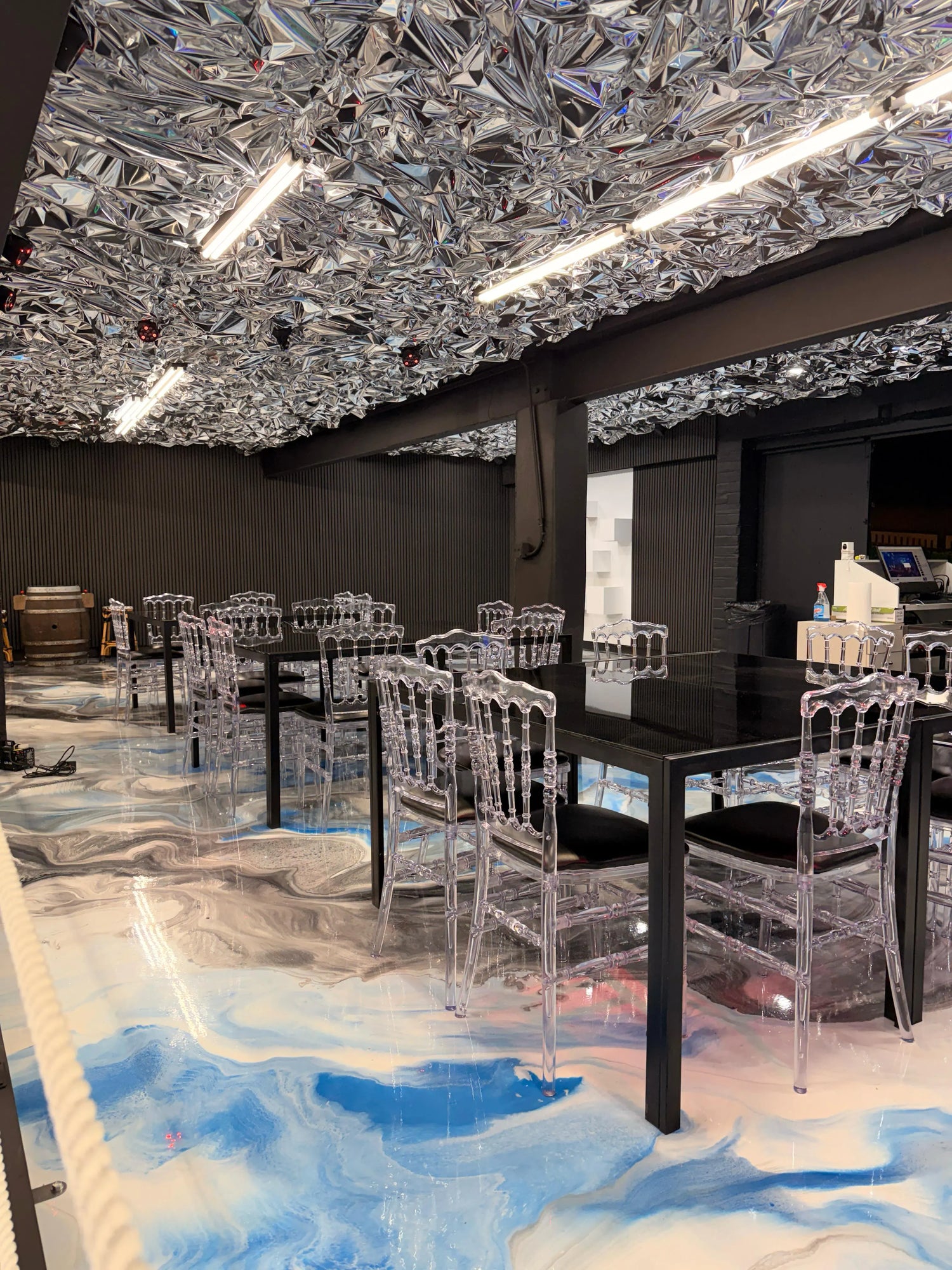 A modern dining area with black tables, transparent chairs, a marbled blue and white floor, and a reflective, crumpled metallic ceiling under bright lights. The space has an artistic, futuristic feel.