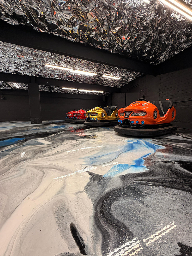 Three bumper cars—two orange and one red—sit on a shiny, marbled floor with black, white, and blue swirls in a dark room with metallic, reflective panels on the ceiling and bright overhead lights.