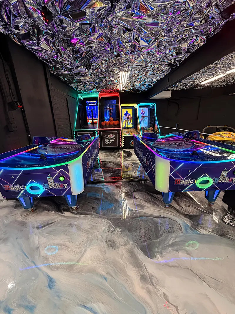 Two air hockey tables with colorful LED lights are in the foreground of an arcade. Behind them are various arcade machines. The ceiling is covered in reflective, metallic material, and the floor has a marbled design.