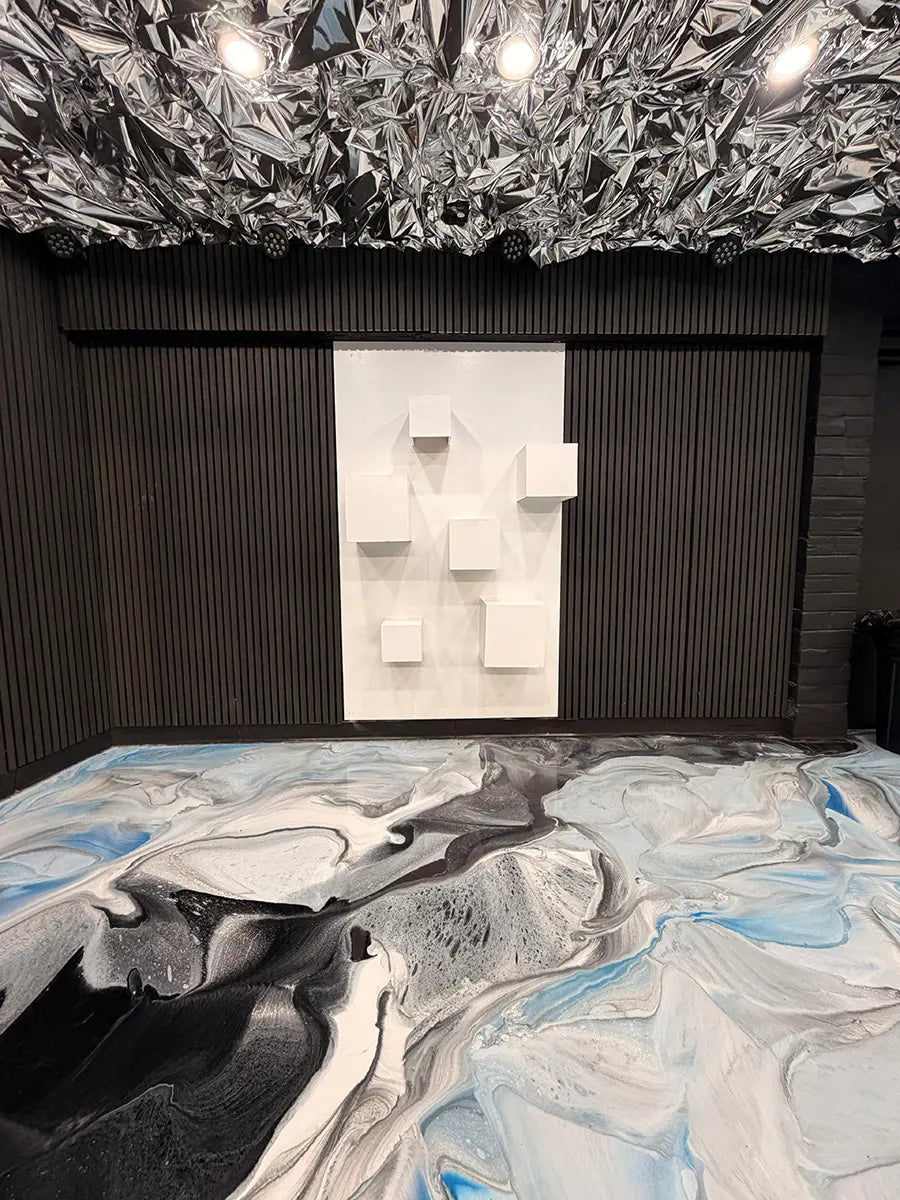 A modern room with a black ridged wall, a ceiling covered in reflective angular panels, abstract black, white, and blue swirled flooring, and a white 3D wall art piece with protruding cubes.