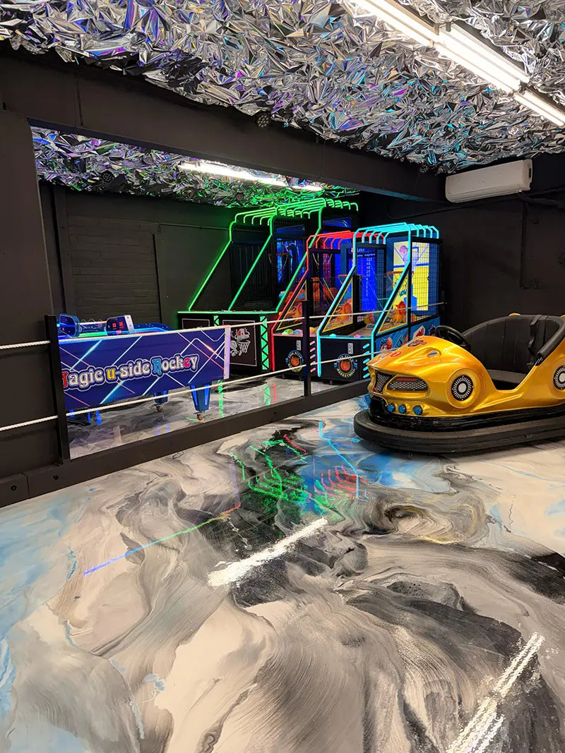 An arcade room with neon-lit game machines, including air hockey and basketball games, next to a yellow bumper car on a shiny, marbled floor with a metallic, reflective ceiling.