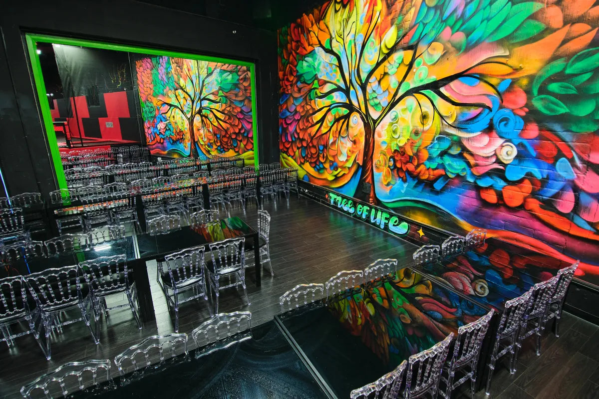 A vibrant room features clear acrylic chairs arranged around black tables, with a large, colorful mural of a tree and flowers labeled "Tree of Life" covering the wall, and a mirror reflecting the mural on one side.