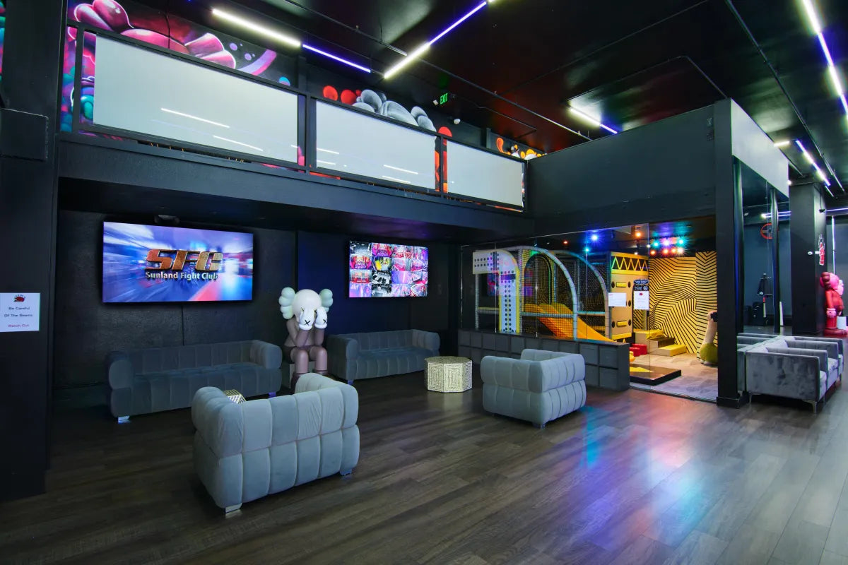 A modern lounge with gray sofas, art sculptures, large TV screens, and neon lighting. A staircase leads to an upper level, and arcade games are visible in a vibrant corner with patterned wall decor.