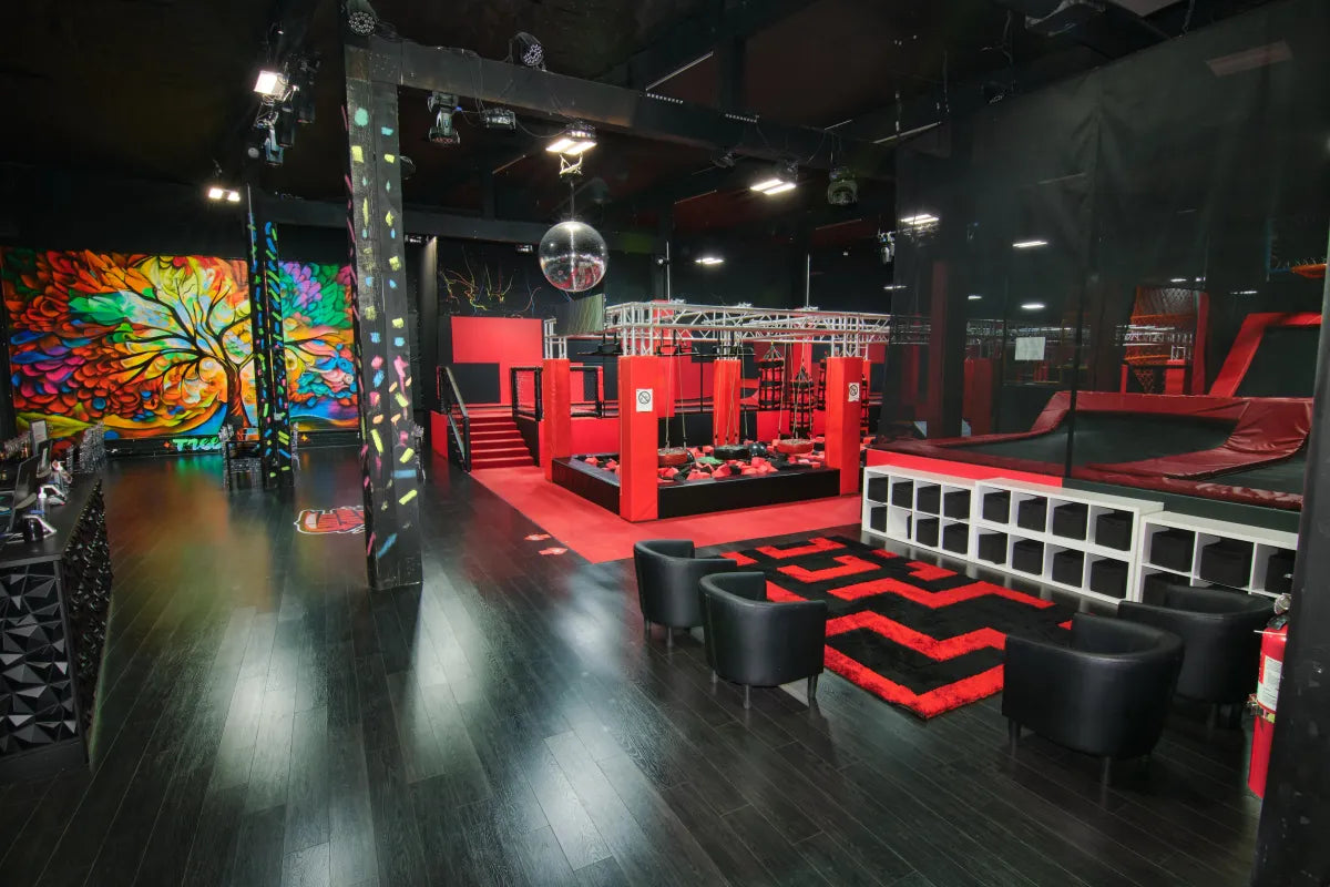 A modern indoor obstacle course gym with red and black decor, padded play structures, a climbing wall, a seating area with black chairs, and a colorful mural of a tree on the wall.