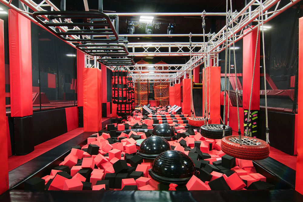 Indoor ninja warrior obstacle course with foam pits, hanging ropes, tire swings, climbing holds, and various equipment, all in red, black, and gray colors, under metal frames and bright overhead lighting.