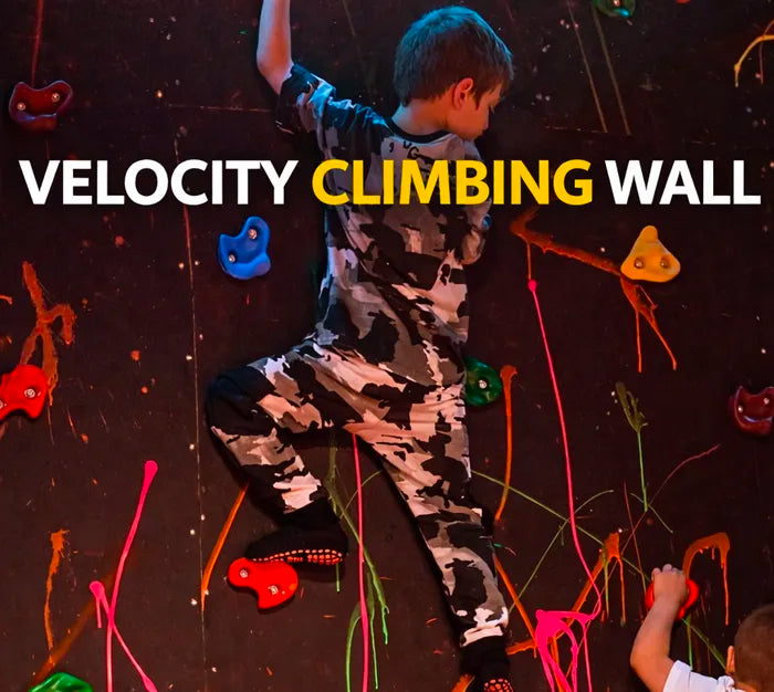 A young child in patterned clothes climbs a colorful indoor climbing wall with bold text reading "VELOCITY CLIMBING WALL" above him. Neon paint splatters and various colored holds decorate the wall.