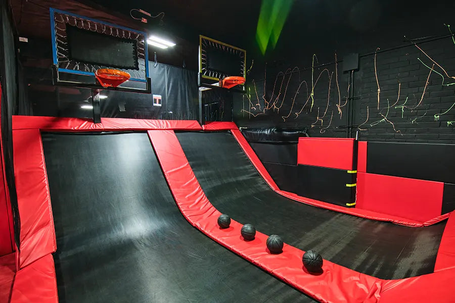 Indoor trampoline basketball court with two hoops, angled trampolines, padded red and black flooring, and four black balls ready for play. Neon lines decorate the dark walls in the background.