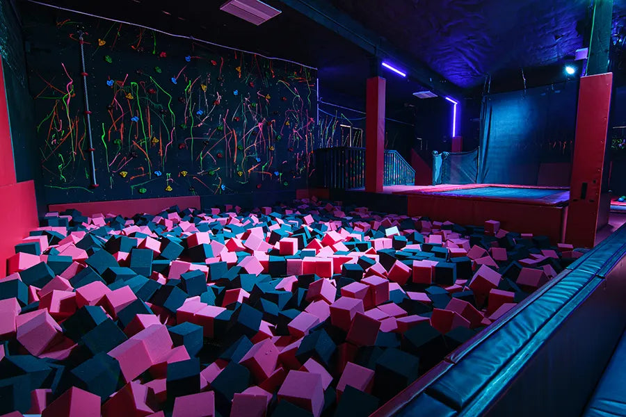 A large indoor trampoline park features a foam pit filled with pink and gray foam cubes, a colorful climbing wall in the background, and glowing purple and blue accent lights.