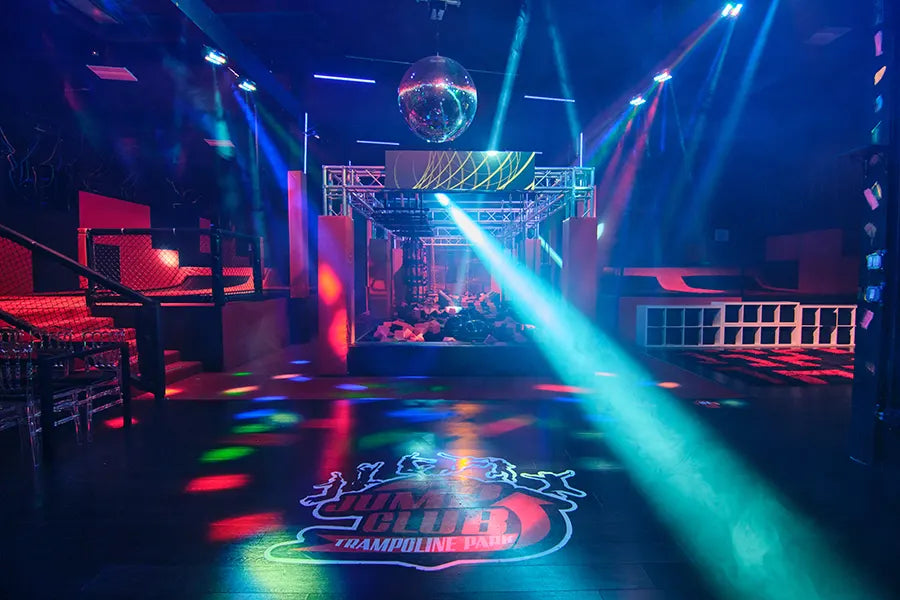 A colorful indoor trampoline park with a disco ball, vibrant stage lights, and a Jump Club logo projected on the dark floor. The area includes foam pits, netted walls, and seating under dynamic, multicolored lighting.