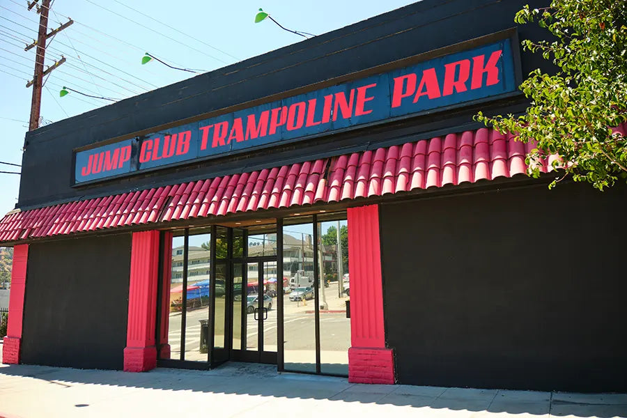 Exterior of a building with a red and blue sign reading "Jump Club Trampoline Park" above the entrance. The facade is black with red pillars and a red-tiled awning. Trees and street are visible in the background.